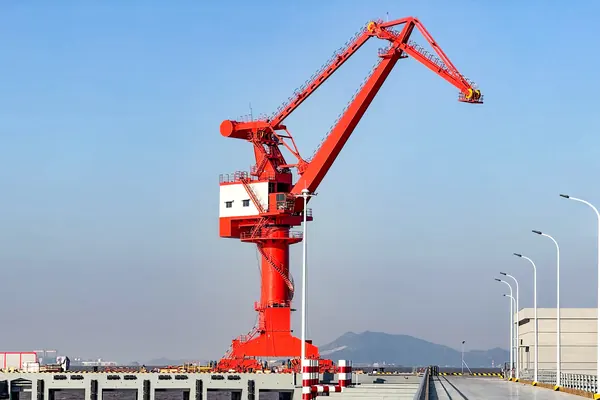 Crane slewing portal berdiri di rel dermaga dengan boom terangkat dan blok kait di ujungnya